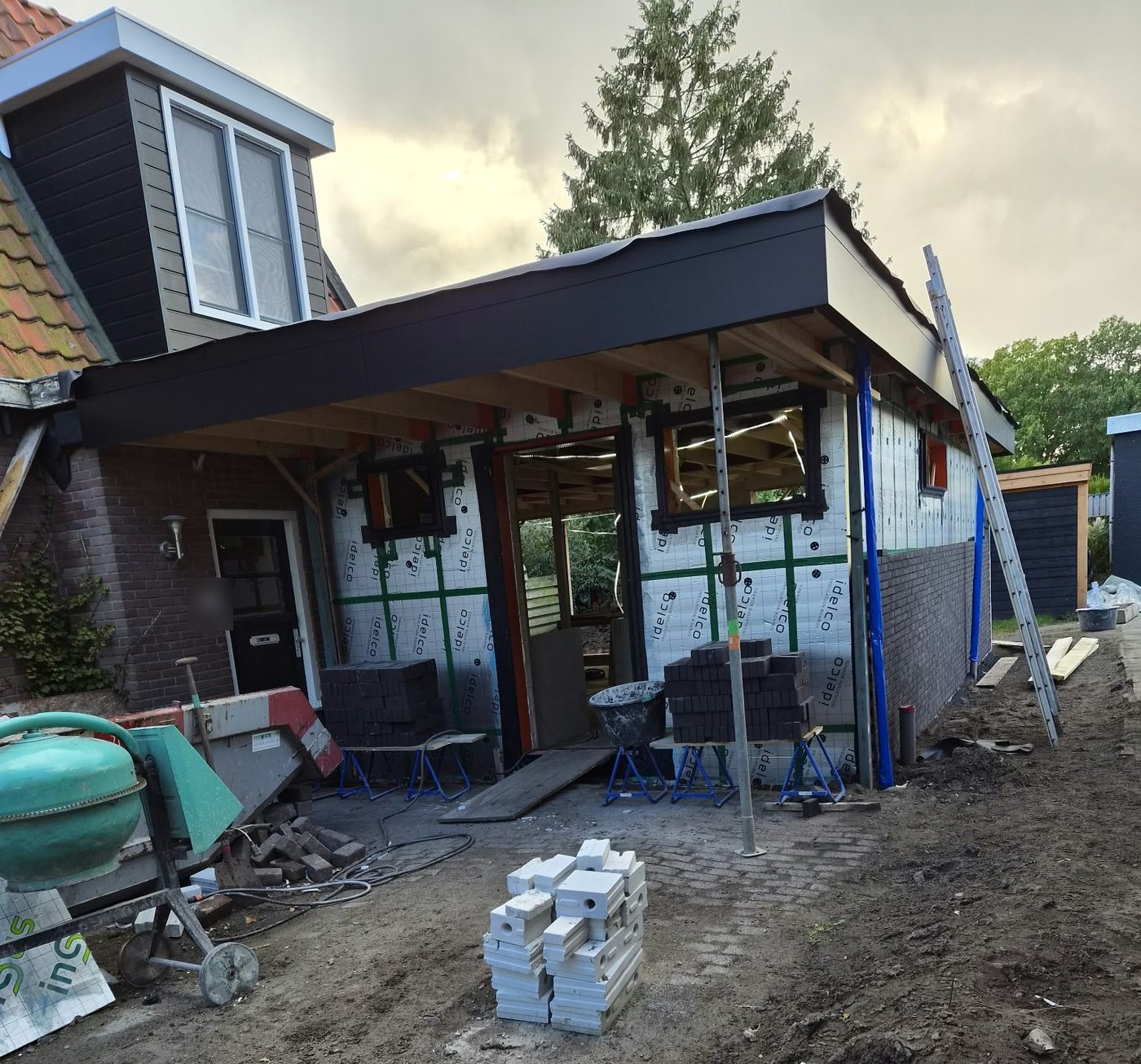 Aanbouw met afgewerkte zwarte dakrand, geïsoleerde gevel en de eerste rijen metselwerk aan de zijkant