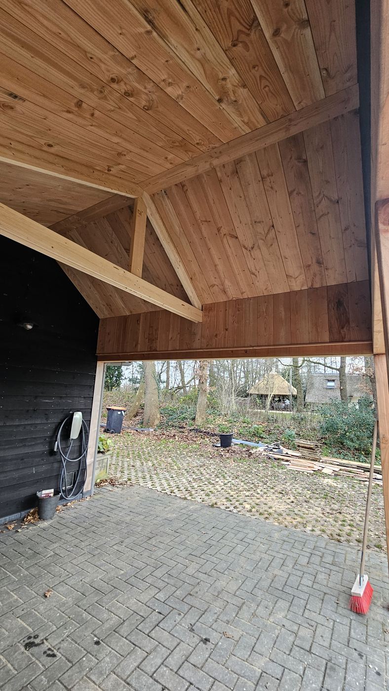 Zicht vanuit de carport naar buiten onder het houten plafond, met zwarte zijwand en klinkervloer