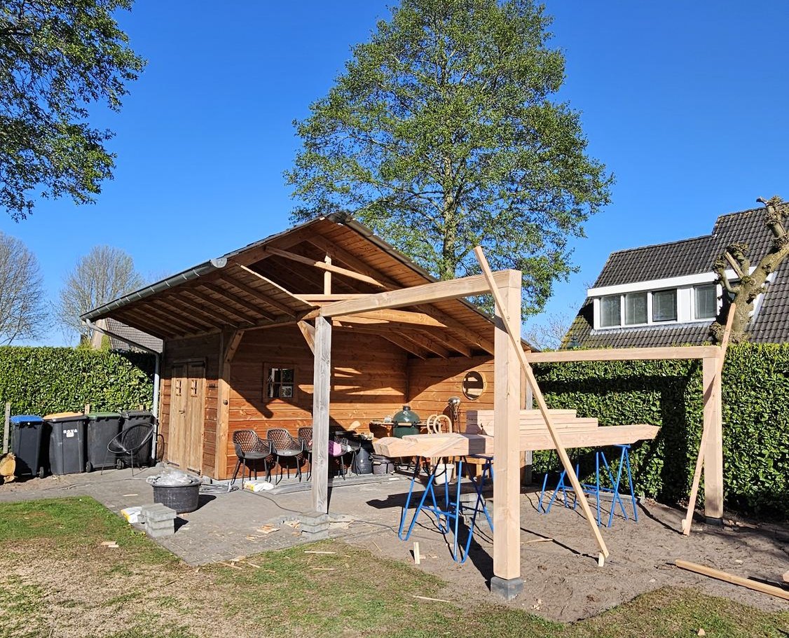 Nieuwe houten staanders en balken naast een bestaand tuinhuis, met schoren voor de opbouw